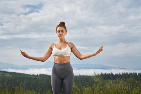 Peaceful caucasian woman in sport clothes standing outdoors with closed eyes and meditating. Beautiful green mountains on background. Spirituality and harmony concept.の写真素材