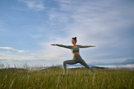 Charming woman in sport clothes doing yoga exercises among summer mountains. Pretty dark haired lady enjoying morning workout on fresh air.の写真素材