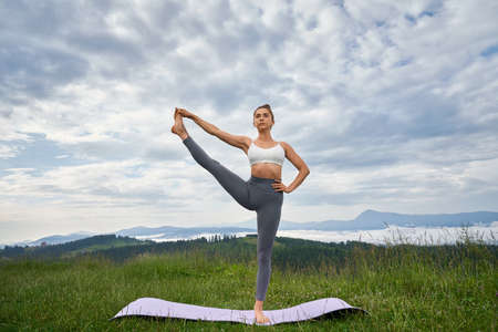 Sporty woman standing on one leg and keeping another outstretched with background of summer mountains. Yoga practice and balancing concept.の写真素材