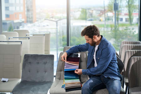 Front view of brunette boy with beard buying furniture in store. Young male sitting, choosing fabric for sofa and chair, holding palette, looking down. Concept of shopping,の写真素材