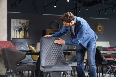 Front view of customer choosing chair in furniture store. Handsome brunette male with beard standing, touching, looking down, buying. Concept of shopping and modern lifestyle.の写真素材