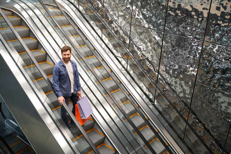 Above view of young male with beard standing, going down by escalator. Stylish brunette man with beard holding bags in shopping mall, smiling. Concept of urban lifestyle.の写真素材