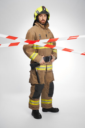 Serious firefighter in full gear standing behind red and white signal tape. Front view of bearded rescuer looking at camera from closed by caution tape area, on white background. Workplace concept.の写真素材