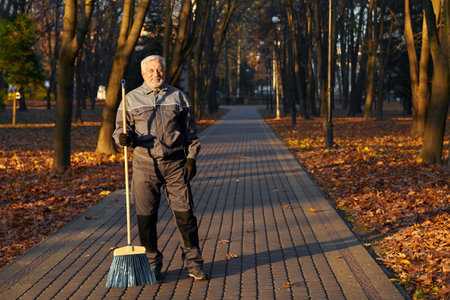 Mature male worker posing to camera with broom in park. Front view of happy bearded man in age, wearing uniform standing with broom and smiling, with park alley on background. Workplace concept.の写真素材