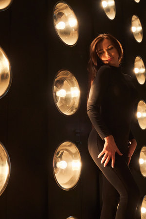 Mysterious, sexy female dancer in black apparel posing with hand on hips in dark room. Side view of...