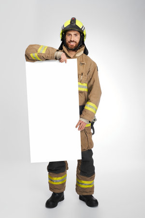 Smiling fireman in yellow helmet standing with blank banner in studio. Front view of bearded firefighter holding placard with copy place, on gray background. Concept of firefighting, announcing.の写真素材