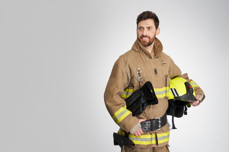 Confident guy in firemans uniform carrying helmet, while posing in studio. Portrait of handsome firefighter looking away and smiling, isolated on gray background. Concept of profession, firefighting.の写真素材