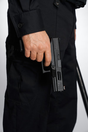 Anonymous man in black uniform carrying short-barrelled handgun, firearm, lowered down. Crop view of policeman with black pistol, holding forefinger on trigger, isolated on white. Concept of weapon.の写真素材