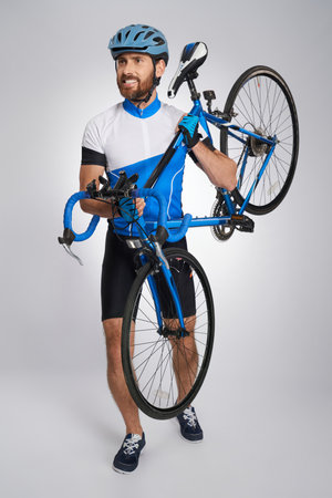 Strong, professional cyclist in helmet carrying bicycle in studio. Front view of handsome bearded athlete in cycle outfit lifting bike, isolated on grey background. Concept of sport activity, cycling.の写真素材
