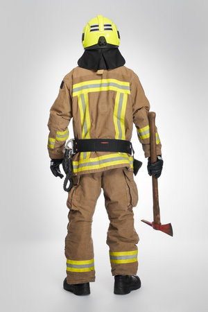 Strong firefighter in helmet and uniform holding fire axe in studio. Back view of unrecognizable male fireman in full gear holding hatchet tool for work ,on gray background. Concept of fire equipment.の写真素材