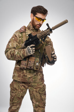 Focused male army soldier in uniform recharging rifle in studio. Portrait of serious man in military apparel reloading weapon, while looking at magazine, on white background. Military weapon concept.の写真素材