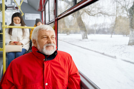 Front view of pensive grizzled male wearing red jacket sitting with passengers behind in bus. Close up of man looking at window while travelling in transport along winter road. Concept of city life.の写真素材