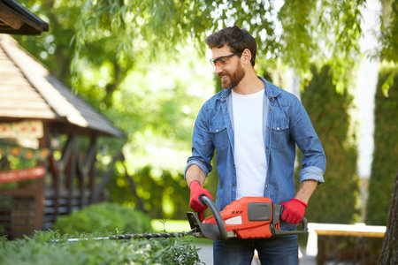 Professional male gardener with beard smiling, while trimming green shrub in garden. Front view of...