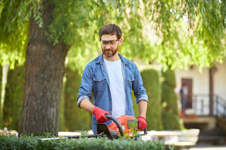 Smiling bearded male gardener cutting boxwood bush with hedge trimmer at sunny day. Front view of...