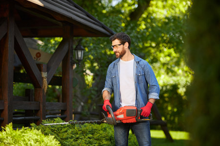 Skilled landscaper in gloves shaping evergreen hedge with trimmer at summer day. Front view of happy man taking care of conifer bush with electric lopper, against wooden gazebo. Concept of gardening.の写真素材