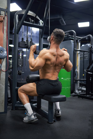 Strong, muscular man pulling training apparatus, while exercising in gym. Back view of anonymous healthy man in shorts, building back muscles, working out. Sport, lifestyle, bodybuilding concept.の写真素材