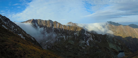 Panoramic view of steep mountains with passes covered with clouds and mountain lake in the valleyの写真素材