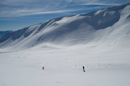 Two hikers move on the snow plateau with steep mountain ridge in the backgroundの写真素材