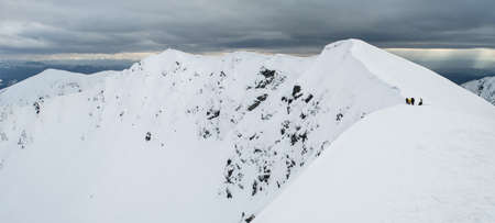 Panoramic view of snowy mountain range with hikers on it in cloudy winter dayの写真素材