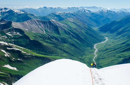 Female climber goes down the snow slope tied to the rope and using the ice ax as a cane. Alpine peaks and mountain valley are in the backgroundの写真素材