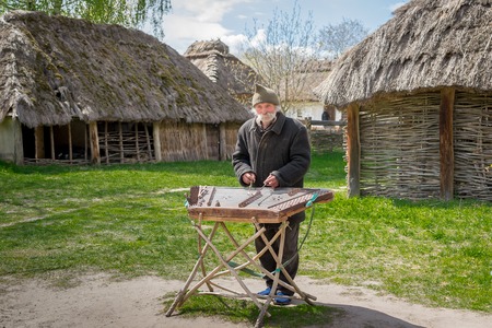 Kyiv, Ukraine - April 23, 2017: An elderly Ukrainian man, a peasant with a long mustache and beard, plays an ancient musical instrument of cymbals. Vintage village with wooden houses. Museum Pyrohivのeditorial素材