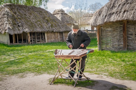 Kyiv, Ukraine - April 23, 2017: An elderly Ukrainian man, a peasant with a long mustache and beard, plays an ancient musical instrument of cymbals. Vintage village with wooden houses. Museum Pyrohivのeditorial素材