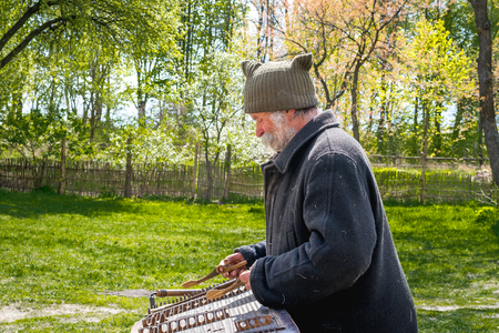 Kyiv, Ukraine - April 23, 2017: An elderly Ukrainian man, a peasant with a long mustache and beard plays an ancient musical instrument of cymbals. Spring landscape. Old Street Musician. Museum Pyrohivのeditorial素材