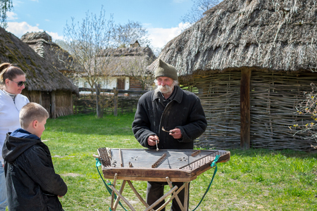 Kyiv, Ukraine - April 23, 2017: An elderly Ukrainian man, a peasant with a long mustache and beard, plays an ancient musical instrument of cymbals. Vintage village with wooden houses. Museum Pyrohivのeditorial素材
