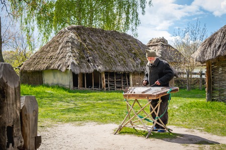 Kyiv, Ukraine - April 23, 2017: An elderly Ukrainian man, a peasant with a long mustache and beard, plays an ancient musical instrument of cymbals. Vintage village with wooden houses. Museum Pyrohivのeditorial素材