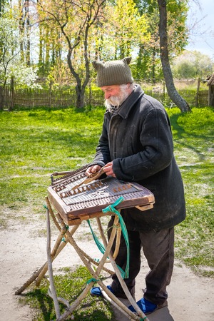 Kyiv, Ukraine - April 23, 2017: An elderly Ukrainian man, a peasant with a long mustache and beard plays an ancient musical instrument of cymbals. Spring landscape. Old Street Musician. Museum Pyrohivのeditorial素材