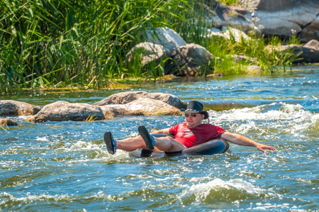 Village Myhiya, Nikolaev region, Ukraine - July 2, 2017: A gentleman in a black hat rafting on a river on a rubber inflatable wheel. A popular place for extreme recreation.のeditorial素材