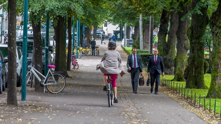 Helsinki, Finland - September 4, 2018. A woman riding a bicycle in a raincoat, pink breeches and a protective helmet in the park. On the way to two men in business suitsのeditorial素材