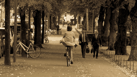 Helsinki, Finland - September 4, 2018. A woman riding a bicycle in a raincoat, pink breeches and a protective helmet in the park. On the way to two men in business suitsのeditorial素材