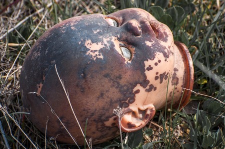 Creepy old doll head. Broken abandoned damaged scary toy in a garbage dump close-up. In the toy, the eye and mouth are damaged. The concept of fear, horror, crime. Very scary.の写真素材