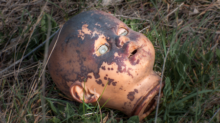 Creepy old doll head. Broken abandoned damaged scary toy in a garbage dump close-up. In the toy, the eye and mouth are damaged. The concept of fear, horror, crime. Very scary.の写真素材