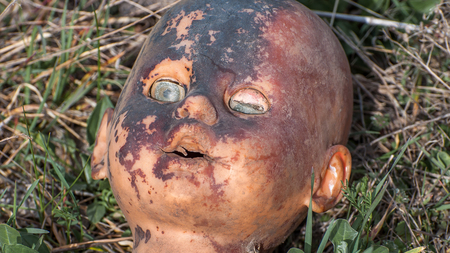 Creepy old doll head. Broken abandoned damaged scary toy in a garbage dump close-up. In the toy, the eye and mouth are damaged. The concept of fear, horror, crime. Very scary.の写真素材