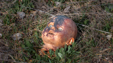 Creepy old doll head. Broken abandoned damaged scary toy in a garbage dump close-up. In the toy, the eye and mouth are damaged. The concept of fear, horror, crime. Very scary.の写真素材