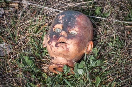 Creepy old doll head. Broken abandoned damaged scary toy in a garbage dump close-up. In the toy, the eye and mouth are damaged. The concept of fear, horror, crime. Very scary.の写真素材