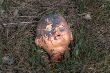 Creepy old doll head. Broken abandoned damaged scary toy in a garbage dump close-up. In the toy, the eye and mouth are damaged. The concept of fear, horror, crime. Very scary.の写真素材