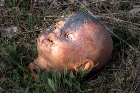 Creepy old doll head. Broken abandoned damaged scary toy in a garbage dump close-up. In the toy, the eye and mouth are damaged. The concept of fear, horror, crime. Very scary.の写真素材
