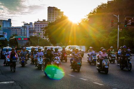 China, Hainan, Sanya - May 13, 2019: Road traffic on the background of the setting sun. Electric scooters - environmentally friendly transport. Glare on the photo.のeditorial素材