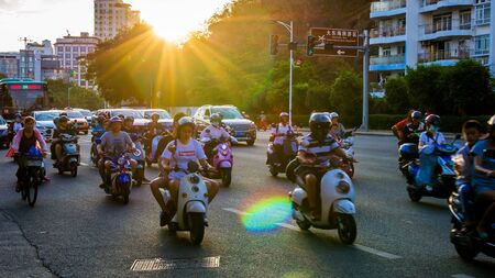 China, Hainan, Sanya - May 13, 2019: Road traffic on the background of the setting sun. Electric scooters - environmentally friendly transport. Glare on the photo.のeditorial素材