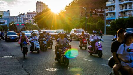 China, Hainan, Sanya - May 13, 2019: Road traffic on the background of the setting sun. Electric scooters - environmentally friendly transport. Glare on the photo.のeditorial素材