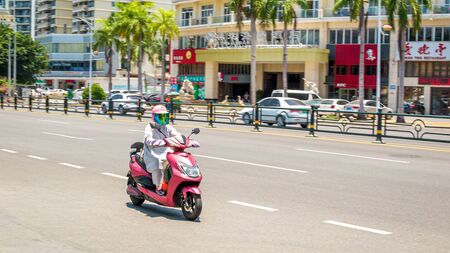 Sanya, Hainan island, China - May 15, 2019: Road traffic. A woman rides an electromoped. 90 percent of mopeds are electric. From 2030, only electric transport will be allowed on the island.のeditorial素材