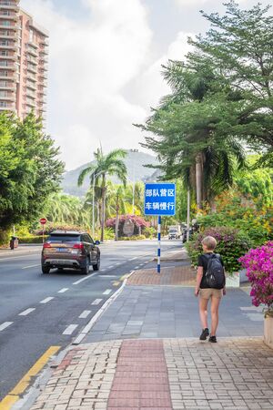 Sanya, Hainan, China - May 16, 2019: a young girl traveler with a backpack is walking along the road, rear view. Along the edges of the road is dense tropical exotic vegetation. Intensive traffic.のeditorial素材