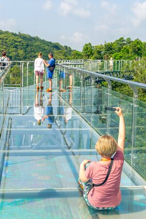 China. The mountains are covered with impassable tropical forests, jungles. Hainan island, Forest Park Yanoda. A tourist girl takes a selfie from a viewing platform in the form of a glass bridge.のeditorial素材