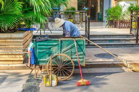 Haikou, Hainan, China - May 12, 2019: A cleaner with a makeshift wagon cleans garbage near the hotel. Broom with bamboo handle. The cart has original wheels.のeditorial素材