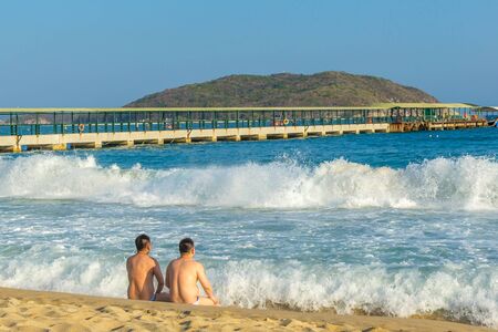 Two men of Asian appearance are sitting on the seashore nearby and are resting, meeting the big waves comingの写真素材