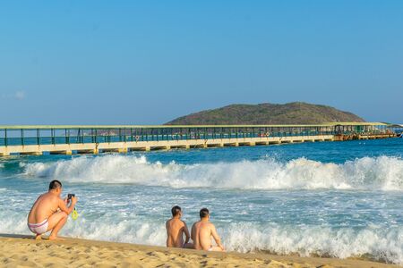Two middle-aged men of Asian appearance are sitting on the seashore nearby and resting, meeting the approaching big waves, and the third is taking pictures of them from behind.の写真素材