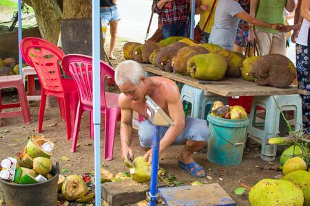 Hainan, China - May 12,2019: A men at a roadside kiosk prepares a coconut with a machete for sale to tourists to drink juice.のeditorial素材